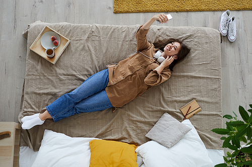 High angle view of happy young woman in casual outfit lying in bed and using smartphone while posing for selfie