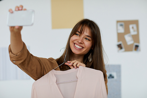 Happy beautiful woman with bangs holding hanger with blouse and taking photo on phone to brag of new blouse to friend