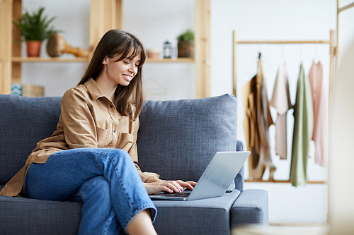 Smiling confident lady entrepreneur in casual outfit sitting on sofa and typing on laptop in own clothing store