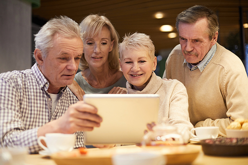 Group of curious elderly former classmates in casual clothing sitting at table and watching old photos on tablet