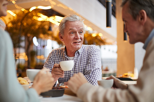 Positive handsome gray-haired mature man in casual shirt sitting at table and talking to friends at tea party in cafe