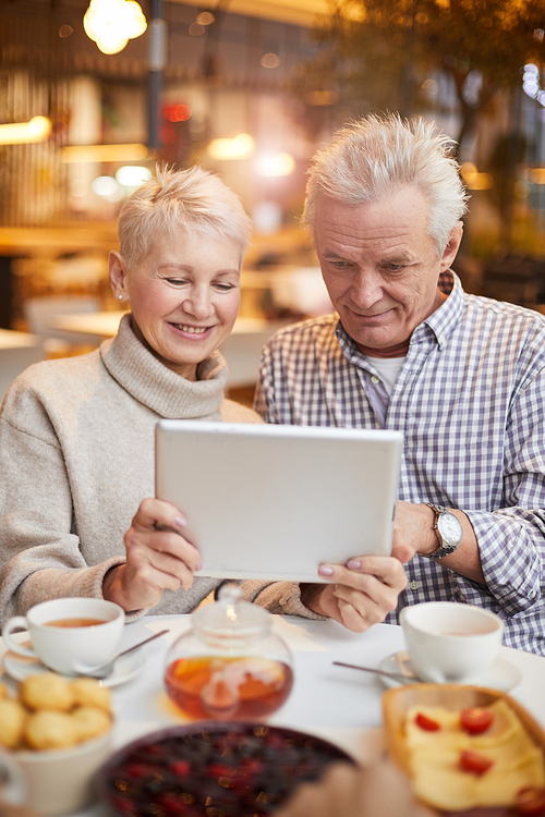 Smiling senior couple in casual outfits sitting at table with tea and desserts and using tablet while watching movie online