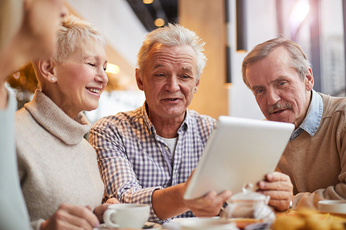 Wrinkled mature man with gray hair sitting at table and using tablet while showing digital photos to friends in cafe