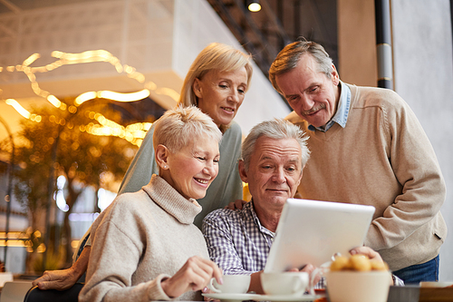 Group of positive senior friends in casual clothing using tablet while surfing websites in cafe with wifi