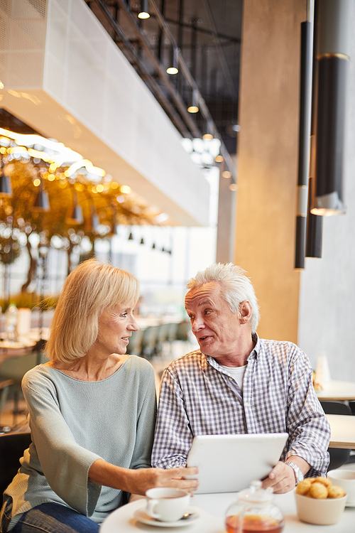 Content modern senior couple in casual outfits sitting at table in restaurant and using tablet while choosing things in online shop