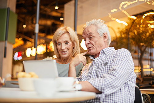 Happy curious senior couple in casual clothing sitting at table and using internet on tablet while spending time in wireless cafe