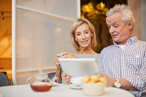 Positive beautiful senior couple sitting at table with tea and sweets and using modern device to watch video