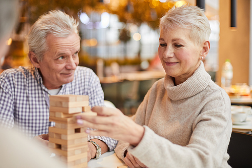 Smiling well-adjusted senior woman in warm sweater taking toy block out of tower while male player staring at her