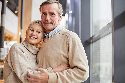 Portrait of happy beautiful senior couple in beige sweaters standing at window and cuddling each other