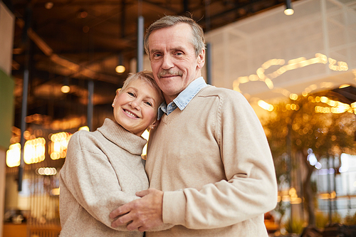 Portrait of happy married elderly couple in casual sweaters embracing each other in modern restaurant full of lights