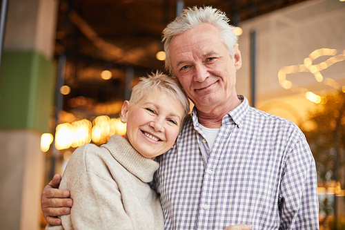 Portrait of positive beautiful elderly spouses in casual outfits hugging each other in modern cafe