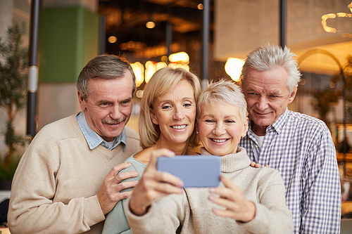 Group of happy senior friends in casual outfits standing in modern cafe and photographing together on smartphone