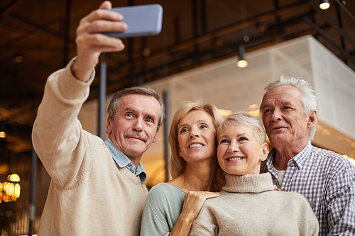 Group of smiling senior friends embracing and posing while man with mustache taking selfie on smartphone