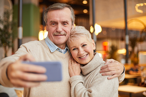 Cheerful beautiful elderly couple in sweaters standing in modern cafe and embracing while taking selfie on phone