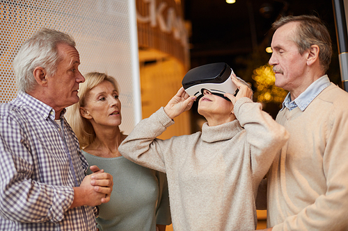 Curious senior woman in sweater looking up while watching 3D movie in VR, her friends looking at her reaction
