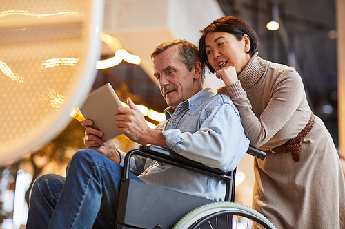 Smiling aged man with mustache sitting in wheelchair and using app on tablet while his Asian wife looking at tablet screen and asking question to him