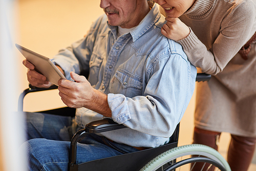 Close-up of positive aged couple watching video on digital tablet: smiling woman leaning on wheelchair of husband