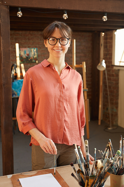 Portrait of young woman standing near the table with brushes and paints and smiling at camera in art studio