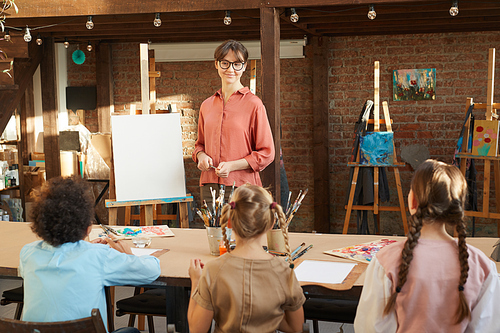 Young female teacher teaching children to paint while they sitting at the table during art lesson in art studio