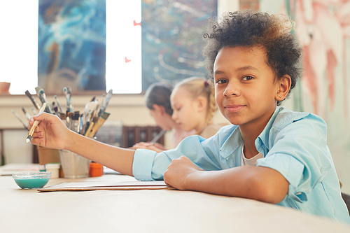 Portrait of African boy looking at camera while sitting at the table and painting during art lesson