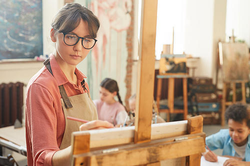 Young teacher in eyeglasses standing near the easel and painting with children sitting in the background in art studio