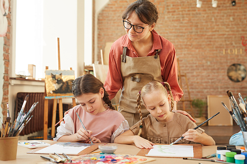 Young teacher in eyeglasses looking at work of little girls who sitting at the table and painting during art lesson at school