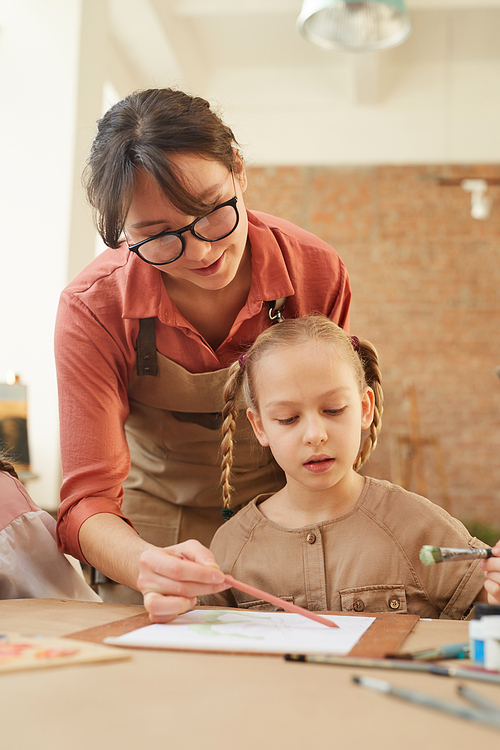 Young woman holding paintbrush and helping little girl to paint during a lesson