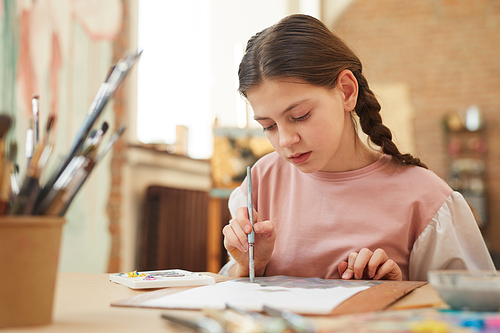 Little girl sitting at the table and painting a picture with brush at the lesson at school