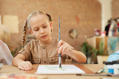 Little girl sitting at the table and drawing a picture with paints and brush in art studio