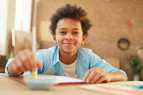 Portrait of African little boy smiling at camera while sitting at the table and painting at the lesson