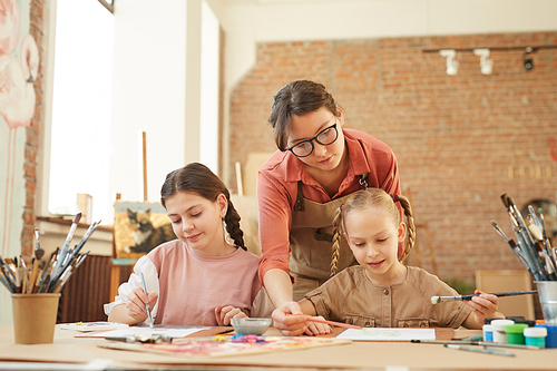 Young teacher teaching her little students to paint a pictures during art lesson at school