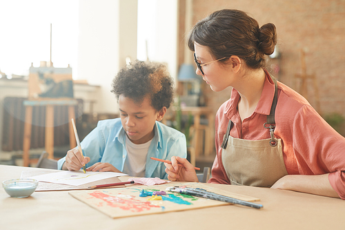 Young teacher sitting at the table together with African schoolboy and teaching him to paint during art lesson