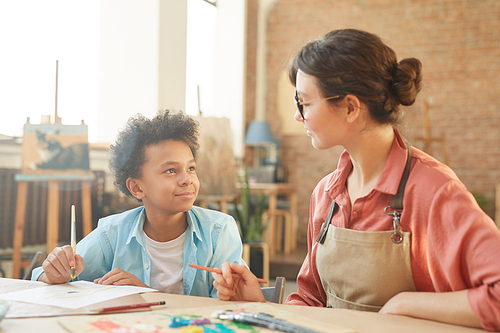 Young woman in eyeglasses talking to African little boy while they sitting at the table and painting during art lesson at school