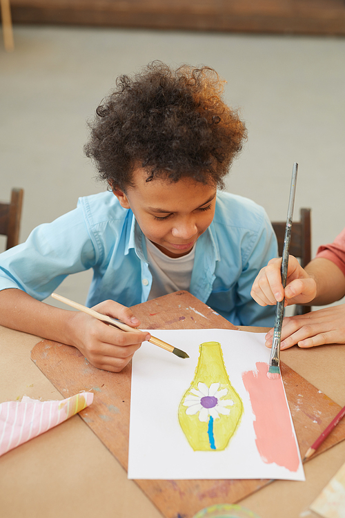 African young boy sitting at the table and painting a picture with paints during art lesson