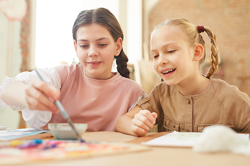 Two happy friends sitting at the table and painting a picture together with paints at school