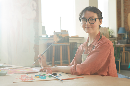 Portrait of young woman in eyeglasses smiling at camera while sitting at the table and using brush and paints at art studio