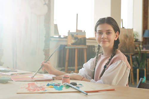 Portrait of little girl looking at camera while painting a picture at the table at art studio