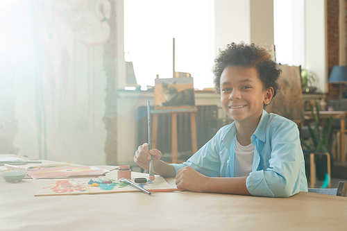 Portrait of African young boy smiling at camera while sitting at the table and drawing a picture with paints and brush