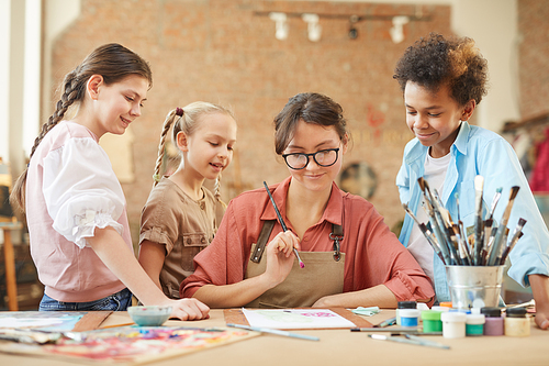 Young teacher in eyeglasses sitting at the table and showing how to draw a picture to her students at art studio