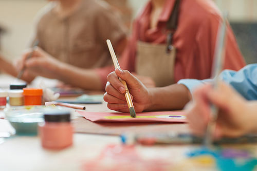 Close-up of child holding paintbrush and painting a picture at the table with other children