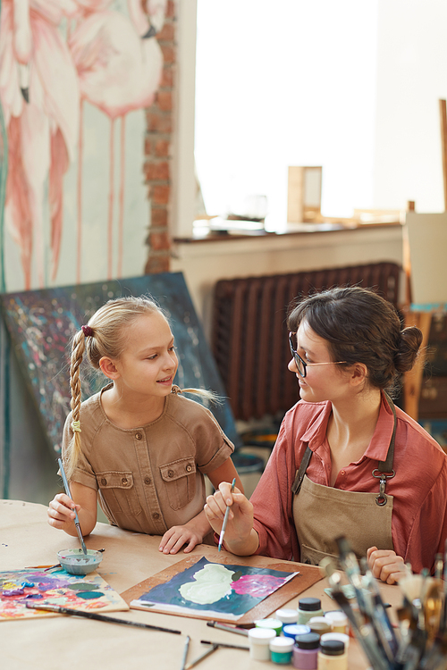 Young woman painting with little girl together at the table and talking at art studio