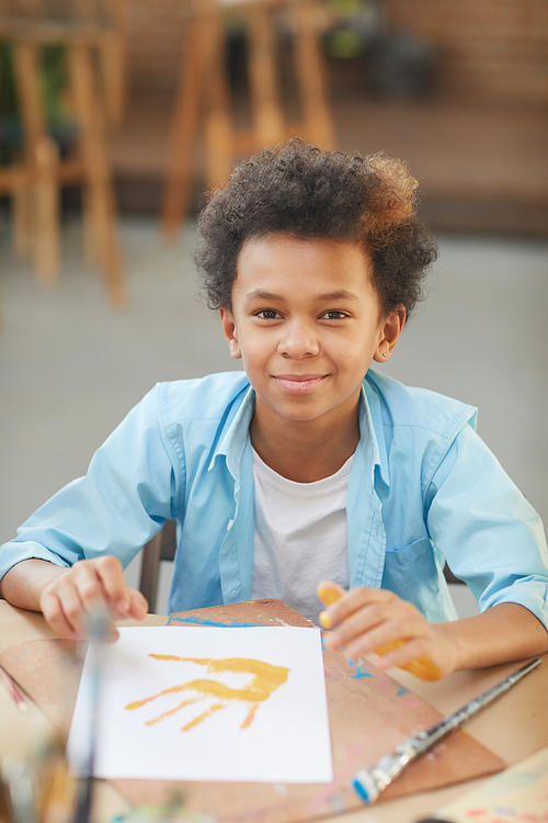 Portrait of African little boy looking at camera while sitting at the table with his handwork in art studio