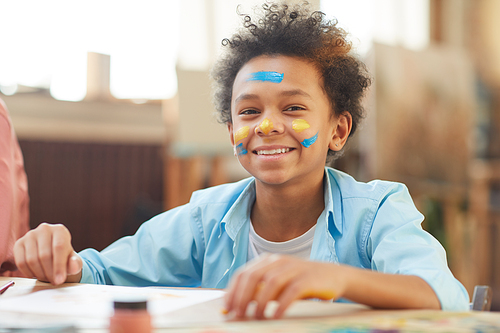 Portrait of African boy with colored paints on his face smiling at camera while sitting at the table at art lesson