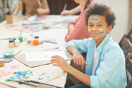 Portrait of African boy looking at camera while sitting at the table with paper and paints at art lesson