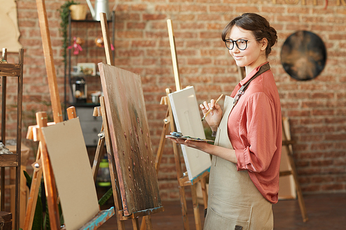 Young artist in eyeglasses and in apron standing in front of easel and she is going to paint in art studio