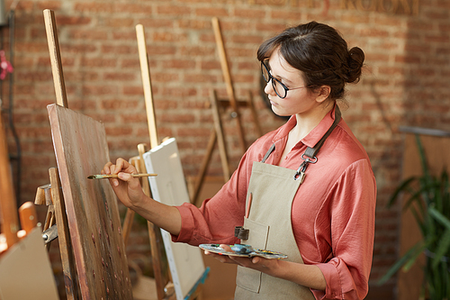 Young woman in eyeglasses painting on easel with paints while standing at art studio