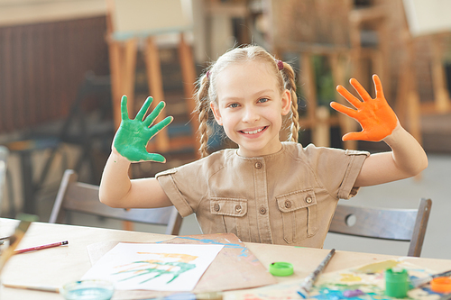 Portrait of happy girl smiling at camera and showing her hands in paints while sitting at the table at art studio