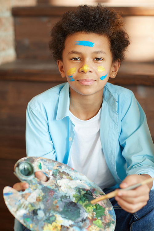 Portrait of African young boy holding palette with paints and looking at camera he painting at studio