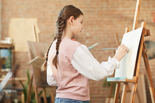 Little girl standing in front of easel and painting on it with paints at art studio