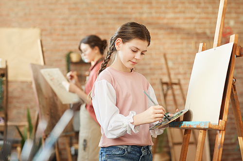 Young girl in casual clothing learning to paint on easel during her lesson at art studio